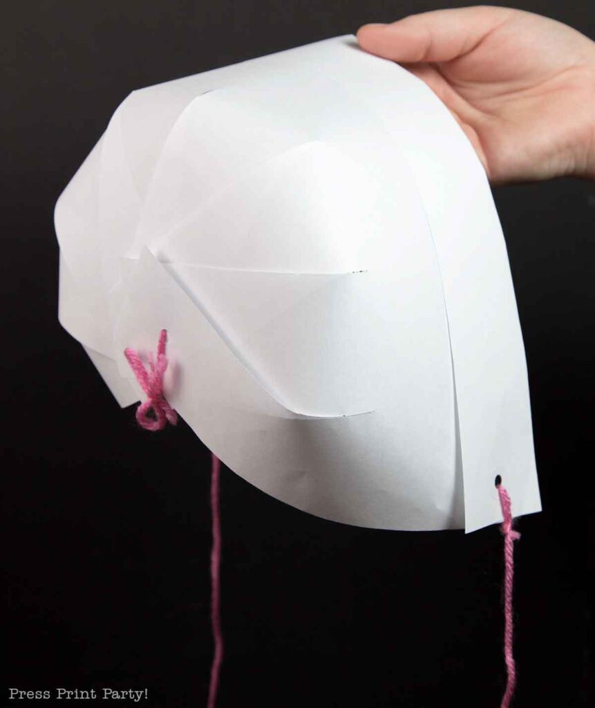 A hand gently holds a whimsical white paper Dodo bird hat, reminiscent of a playful pilgrim bonnet, complete with charming pink yarn ties.