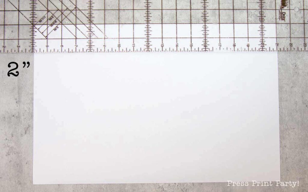 A blank sheet of white paper rests on a cutting mat with measurement markings, including a visible 2-inch label and various rulers. A sketched pilgrim bonnet and the text "Press Print Party!" adorn the bottom right corner, adding a whimsical touch to the scene.