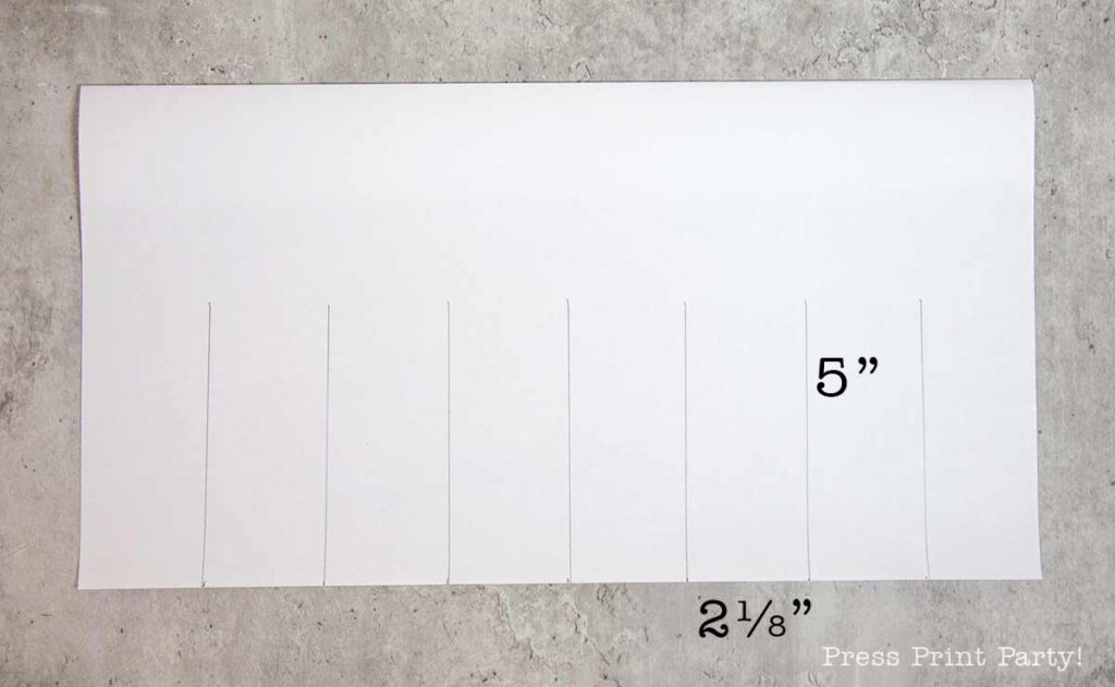 A white sheet of paper with six vertical slits partially cut from the bottom edge evokes the simplicity of a pilgrim bonnet. The paper, marked with measurements of "5 inches" in height and "2 1/8 inches" in width, rests on a textured gray background.