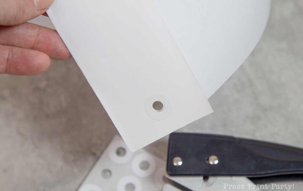 A hand holds a piece of white paper with a circular hole punch. Below are a black hole punch and sticky reinforcement labels, reminiscent of a pilgrim bonnet's neat simplicity. The background is a light gray surface.