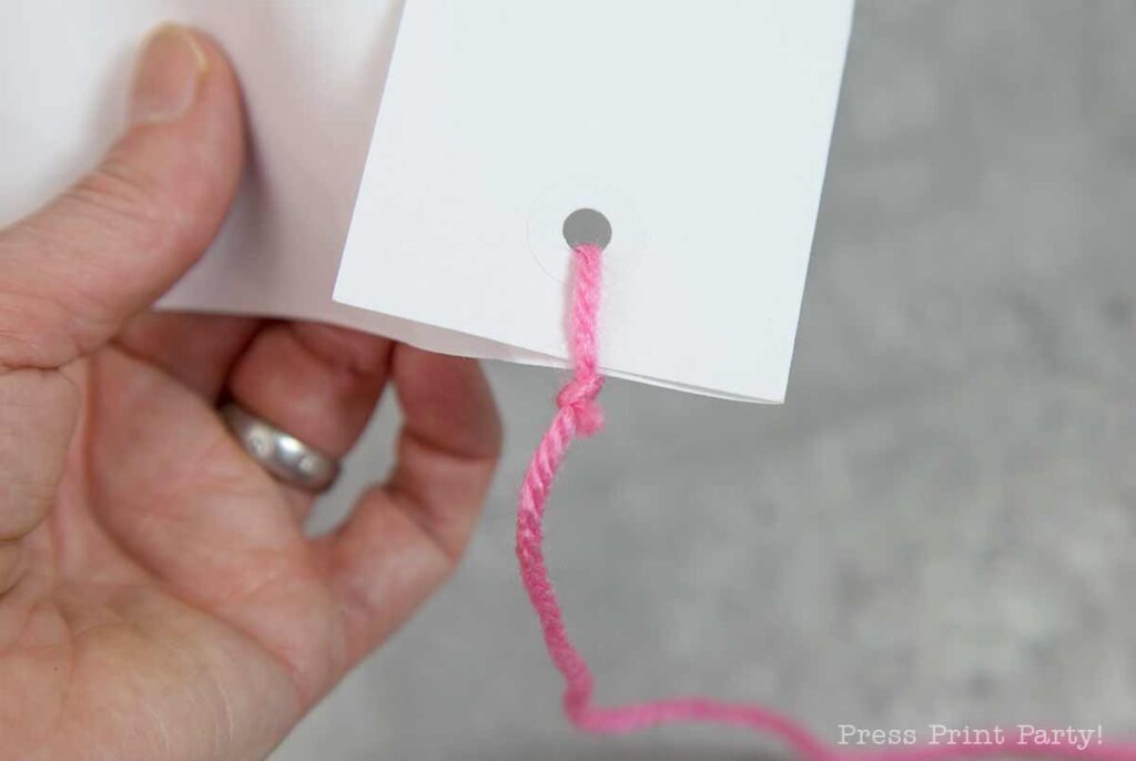 A close-up of a hand holding a piece of white paper with a hole punched at one corner. A pink yarn, reminiscent of a pilgrim bonnet tie, is threaded through the hole and ends in a knot. The background is a soft gray surface.