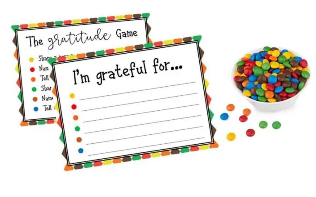 Two Gratitude Game cards with colorful borders, featuring prompts like "I'm grateful for..." and associated actions. A bowl of colorful candies is placed beside the cards, with some candies scattered around.