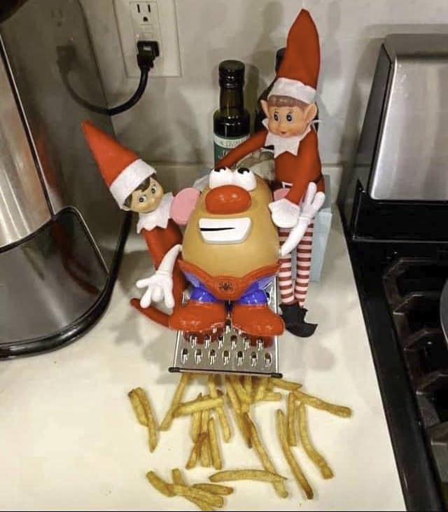 Three toy elves are gathered on a kitchen counter. One elf holds a grater while a Mr. Potato Head toy sits on it, with toy fries scattered below. A bottle of olive oil and other appliances are in the background.