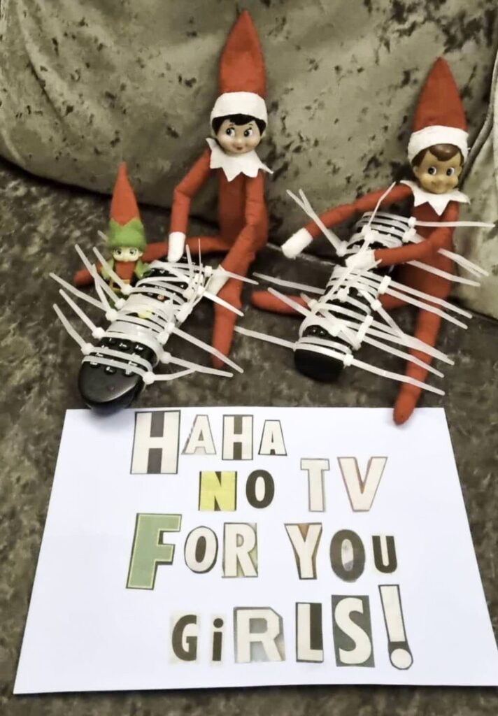 Three elf dolls with red outfits sit on a couch, looking like mischievous versions of the classic bad elf on the shelf. Two are holding a remote control wrapped in zip ties, while a small green elf sits nearby. In front, a sign humorously declares, "HAHA NO TV FOR YOU GIRLS!" in colorful letters.
