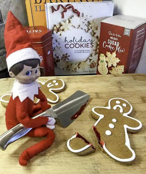 A toy elf in a red outfit sits on a kitchen counter next to holiday cookie boxes. It holds a large silver knife, appearing to have cut a gingerbread cookie into pieces. Gingerbread crumbs are scattered around.
