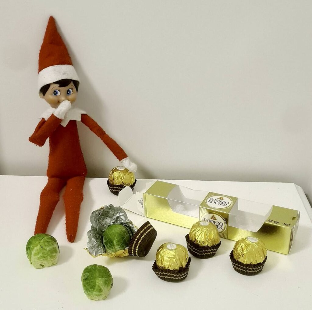 A bad elf on the shelf in a red outfit sits beside a partially open box of chocolates. Three chocolates are wrapped in gold foil, while four brussels sprouts mischievously mimic their appearance, adding a cheeky twist to the scene.