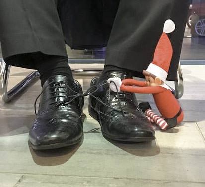 A mischievous elf toy in a Santa hat ties together the shoelaces of a person wearing shiny black shoes and black pants, sitting on a chair.