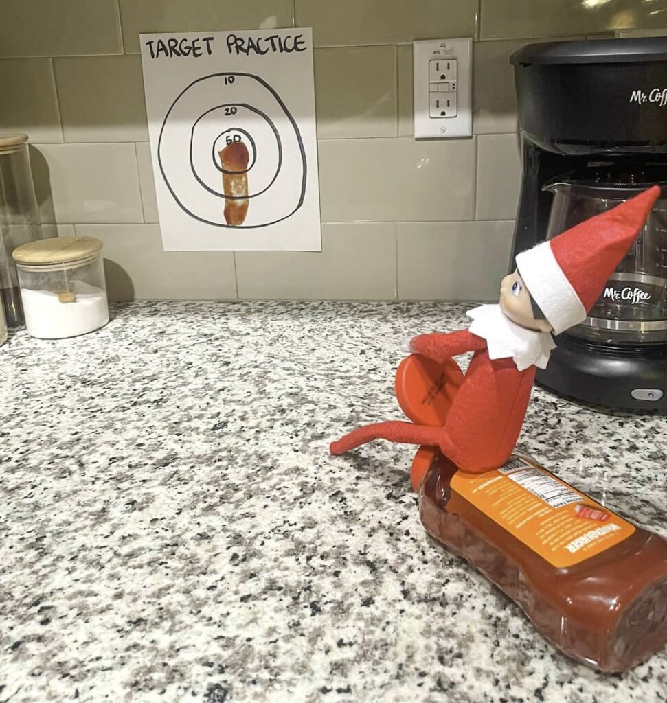 A toy elf is sitting on a bottle of hot sauce on a kitchen counter, aiming a lid like a frisbee at a target practice sheet on the wall. The counter has a coffee maker and a jar in the background.