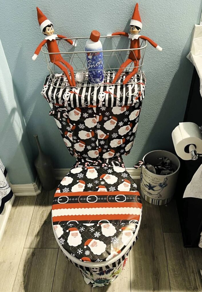 Two bad elf figures sit in a basket above a toilet, wrapped in festive Christmas gift paper with Santa and snowflake designs. A can labeled "Snow" is nestled between them, creating a mischievous holiday-themed setup in the bathroom.
