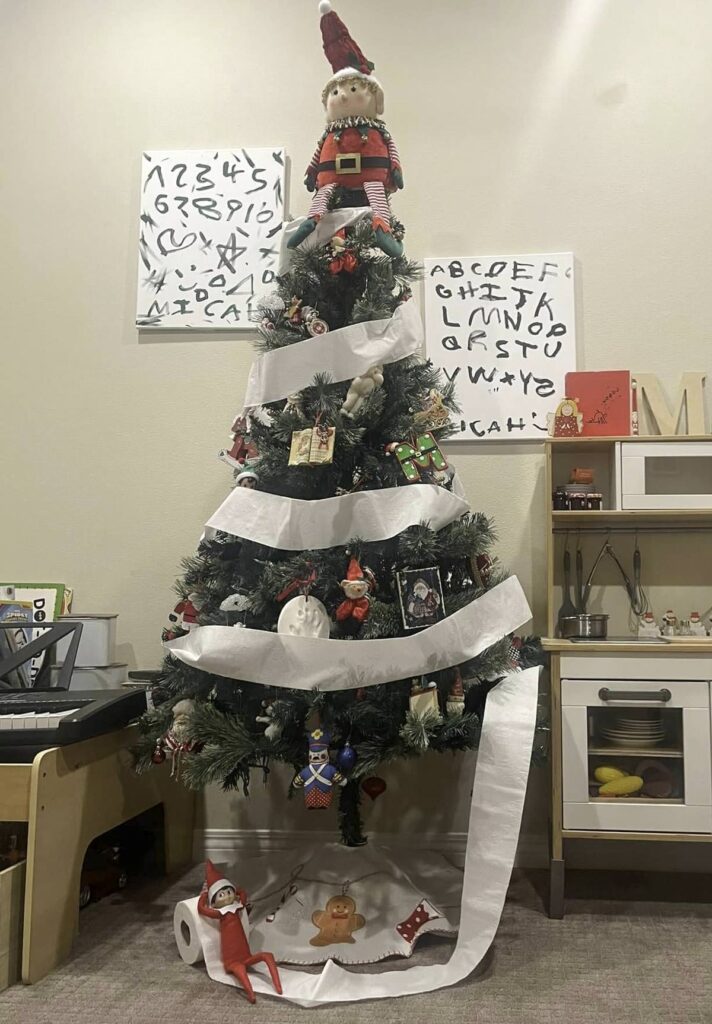 A decorated Christmas tree with white ribbon and various ornaments stands in a cozy room. A plush elf sits atop the tree, while a toy elf and gingerbread figure rest beneath it. A small play kitchen and artwork with letters are in the background.