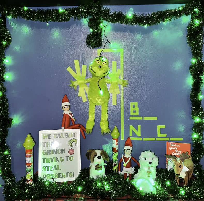 A festive decoration featuring a Grinch plush hanging among glowing green lights, with elves, stuffed dogs, and a sign that reads "We caught the Grinch trying to steal presents!" A "How the Grinch Stole Christmas" book is also displayed.