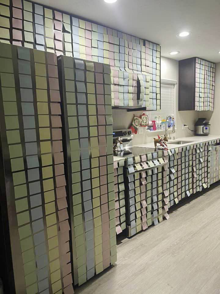 A kitchen covered in multicolored sticky notes resembles a mischievous scene, as if a bad elf on the shelf had decided to leave its playful mark. Cabinets, walls, and appliances, like the fridge and oven, are entirely concealed with sticky notes in shades of green, blue, pink, and yellow.