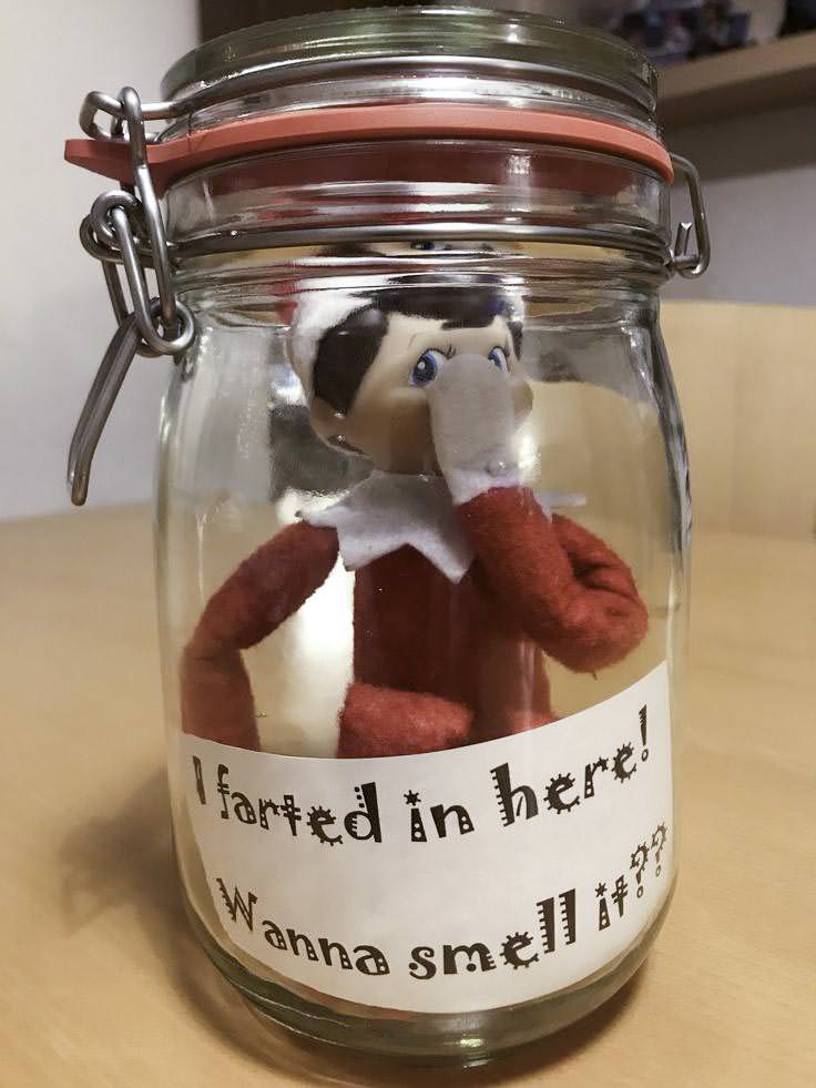 A toy elf is trapped inside a glass jar with its hand covering its nose. A playful sign on the jar reads, "I farted in here! Wanna smell it?!" The jar has a metal latch and is on a wooden surface.