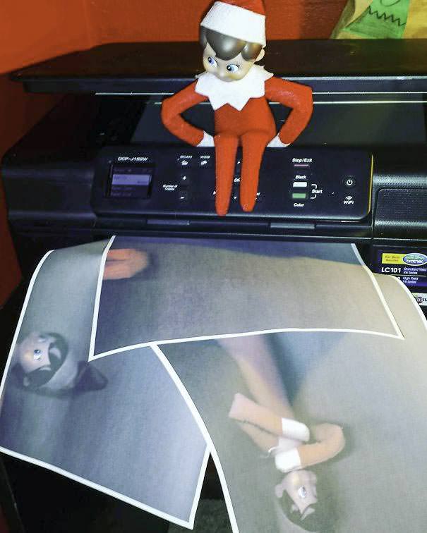 An elf doll sits on a printer while a sheet of paper with its images is being printed. The elf wears a red outfit with a white collar and hat. The printout shows multiple distorted images of the elf.