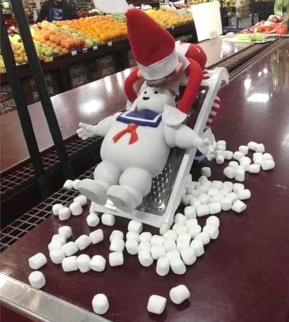 An elf figure is pressing a white marshmallow man toy onto a cheese grater, surrounded by scattered marshmallows, set on a grocery store counter.