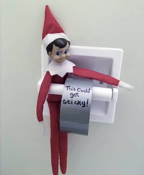 A mischievous elf sits on a toilet paper holder, grasping a roll of duct tape instead of toilet paper. With "This could get sticky!" scrawled in black ink, this bad elf on the shelf dons a red and white outfit with a pointed hat.