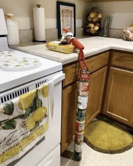 An elf doll is perched on a kitchen counter next to a box of crackers. It appears to have climbed a tower of stacked canned goods to reach the counter. The kitchen is decorated with lemon-themed items and has various fruits displayed.