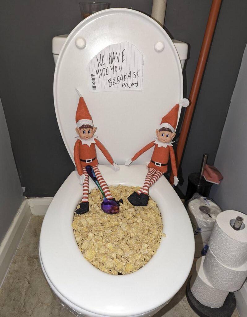 Two mischievous elf dolls, part of the notorious bad elf on the shelf gang, sit on a toilet seat filled with cereal flakes. A cheeky note on the lid reads, "We have made you breakfast." Nearby, toilet paper rolls are precariously stacked.