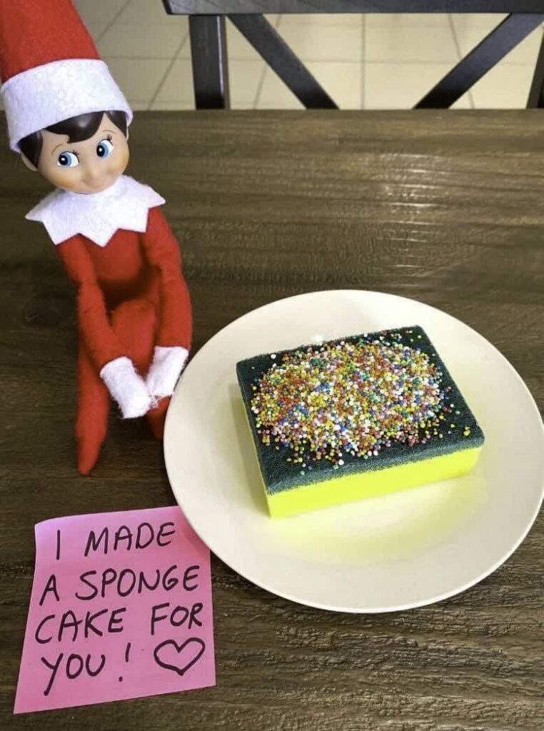 A toy elf sits on a table next to a plate with a sponge covered in colorful sprinkles. A pink note reads, "I made a sponge cake for you!" with a heart symbol.