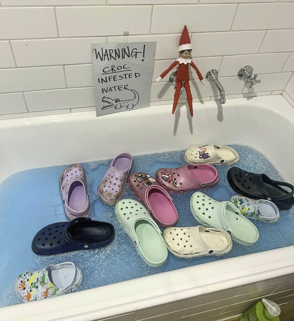 A bathtub filled with blue water and several colorful Crocs floating. A sign above warns "Croc infested water" with a drawing of a crocodile. An elf toy sits on the edge of the tub.