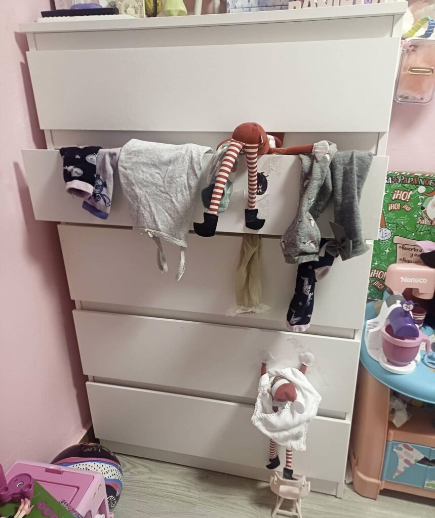 A white dresser in a child's room has drawers slightly open with plush toys, like a mischievous elf on the shelf, hanging over the edges. The room contains various children's toys and items with colorful decorations on the walls.