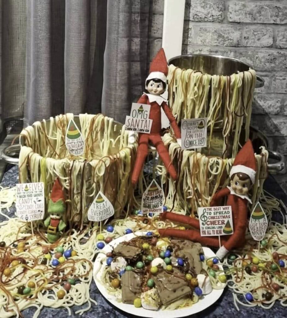 Elves, much like a bad Elf on the Shelf, sit among pots overflowing with spaghetti and syrup, reminiscent of a scene from the movie "Elf." Cookies topped with syrup and candy are on a plate. Signage around them features spirited holiday messages.