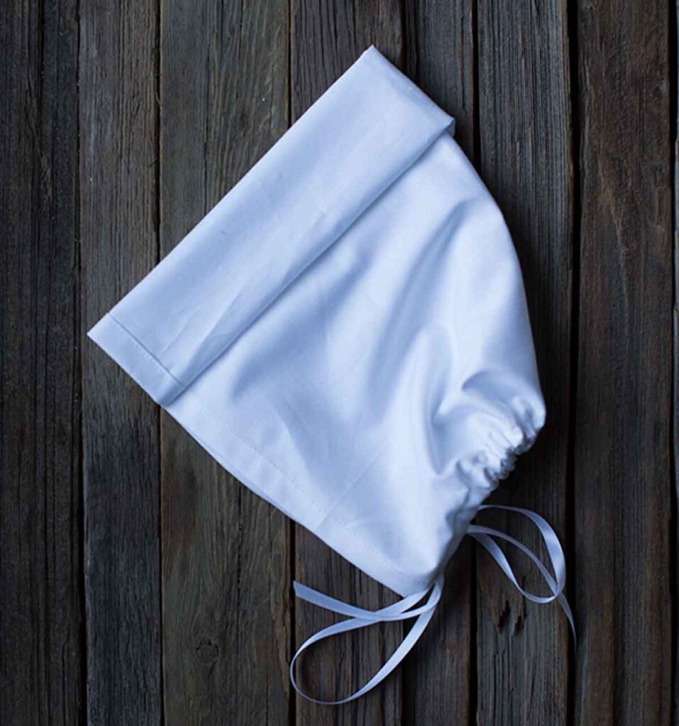 A white bonnet with a folded brim and tied strings rests against a background of weathered wooden planks.