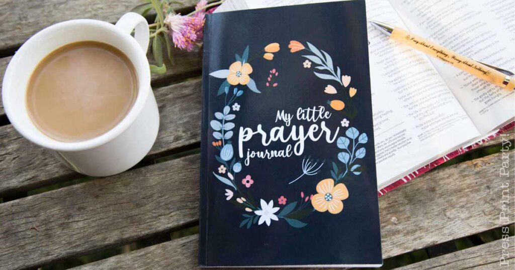 A wooden table with a cup of coffee, an open book with a pen, and a dark-colored notebook titled "My Little Prayer Journal," adorned with floral illustrations.