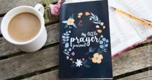 A wooden table with a cup of coffee, an open book with a pen, and a dark-colored notebook titled "My Little Prayer Journal," adorned with floral illustrations.