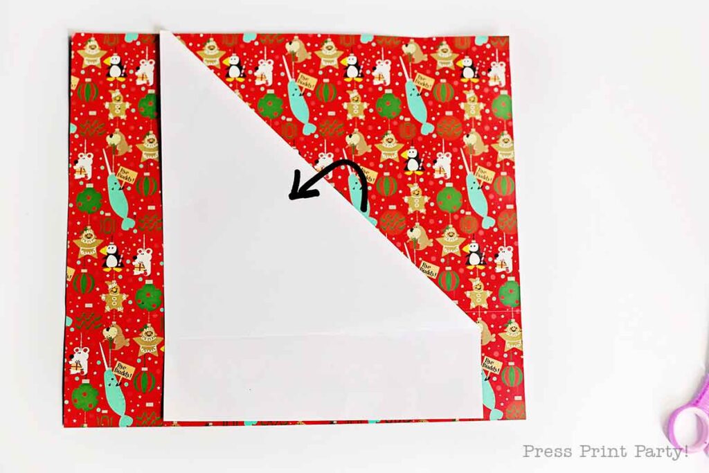 A sheet of festive red gift wrap with Christmas-themed designs is partially folded diagonally. An arrow drawn on the white side indicates the folding direction. A pair of pink scissors is partially visible in the corner.