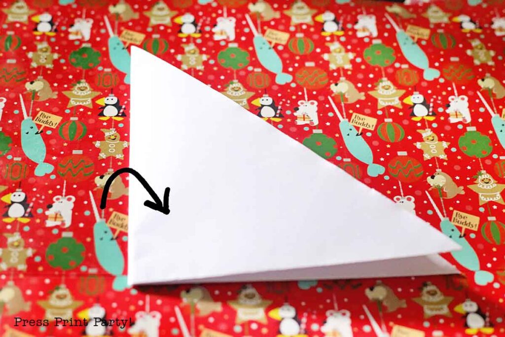 A piece of white paper folded into a triangle on top of red Christmas-themed wrapping paper with festive illustrations, including ornaments, polar bears, and trees. A black arrow is drawn on the paper, pointing to a corner.