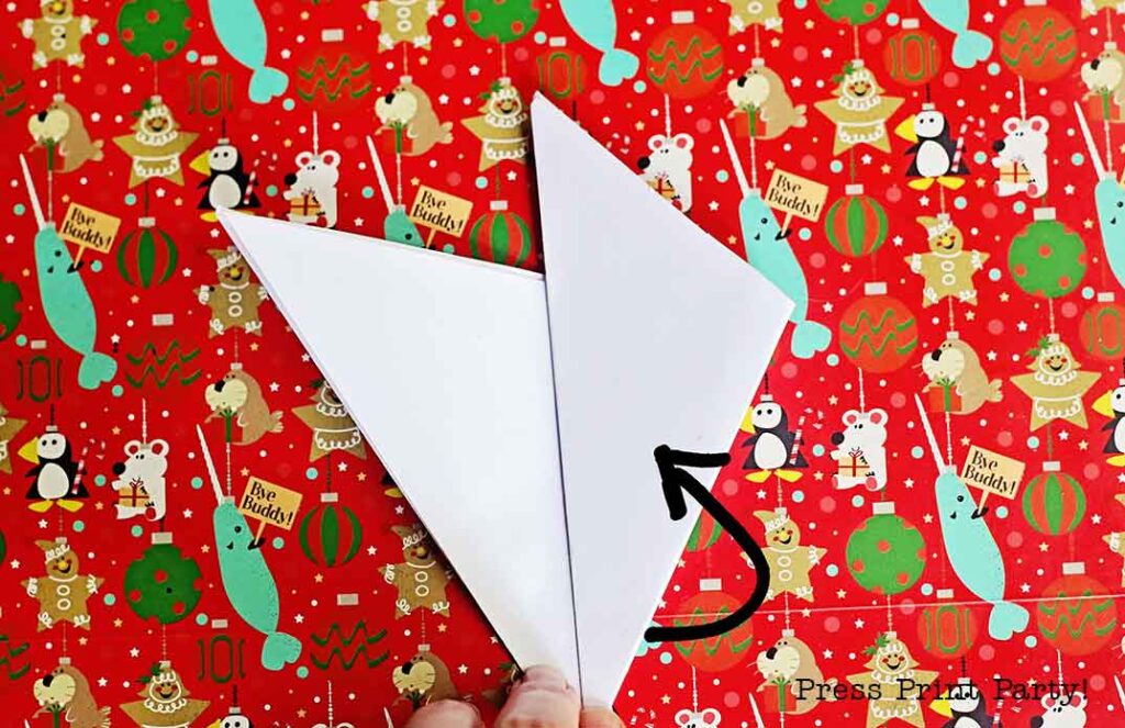 A piece of white paper is partially folded on a colorful holiday-themed background with penguins, narwhals, and ornaments. An arrow is drawn on the paper, indicating the folding direction.