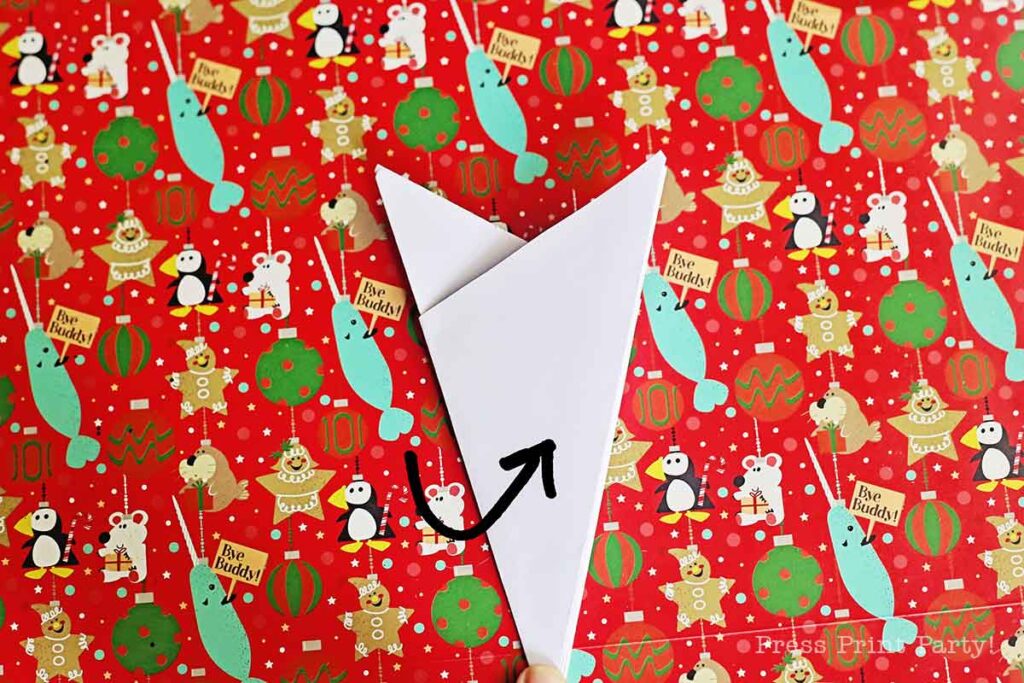 A red Christmas-themed wrapping paper featuring animals like bears, narwhals, and birds. The paper is adorned with festive decorations and text. A white triangle with a black arrow is placed on top, pointing upwards.