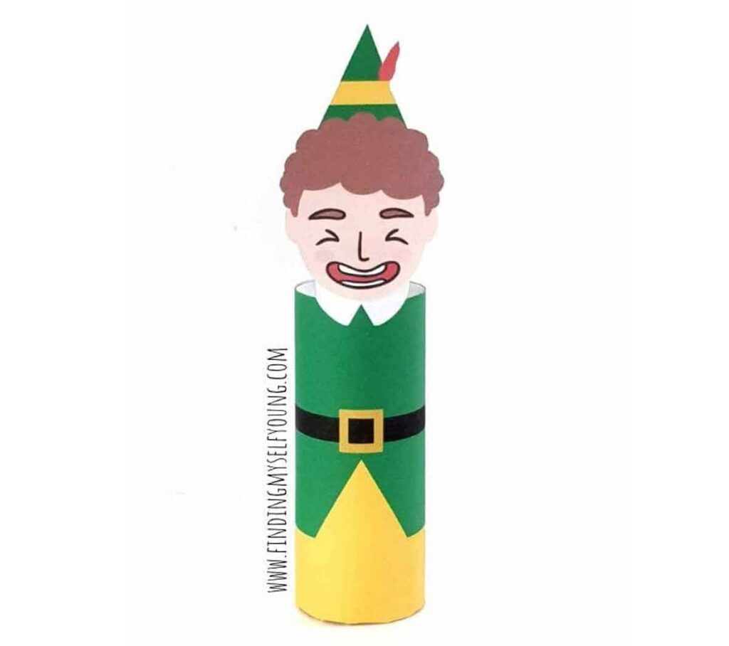 A whimsical elf figure made from a cardboard roll and paper. The elf has a smiling face with curly hair, wearing a green and yellow outfit with a pointed hat adorned with a red feather.