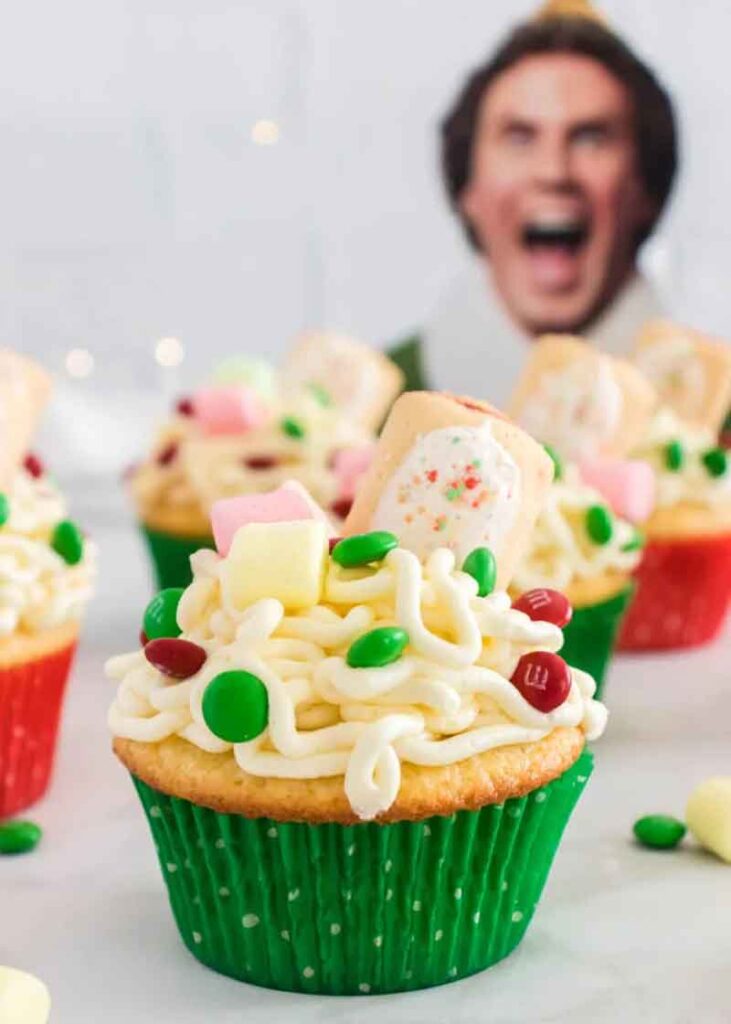 Festive cupcakes topped with creamy white frosting, colorful candies, and small marshmallows. A cutout of a grinning person's face is blurred in the background, adding a playful touch to the holiday-themed scene.
