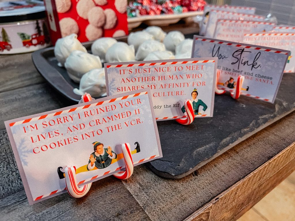 A display of holiday-themed cards held by small candy canes on a slate surface. Each card features humorous quotes. In the background, trays filled with confections are visible, adding a festive touch to the scene.