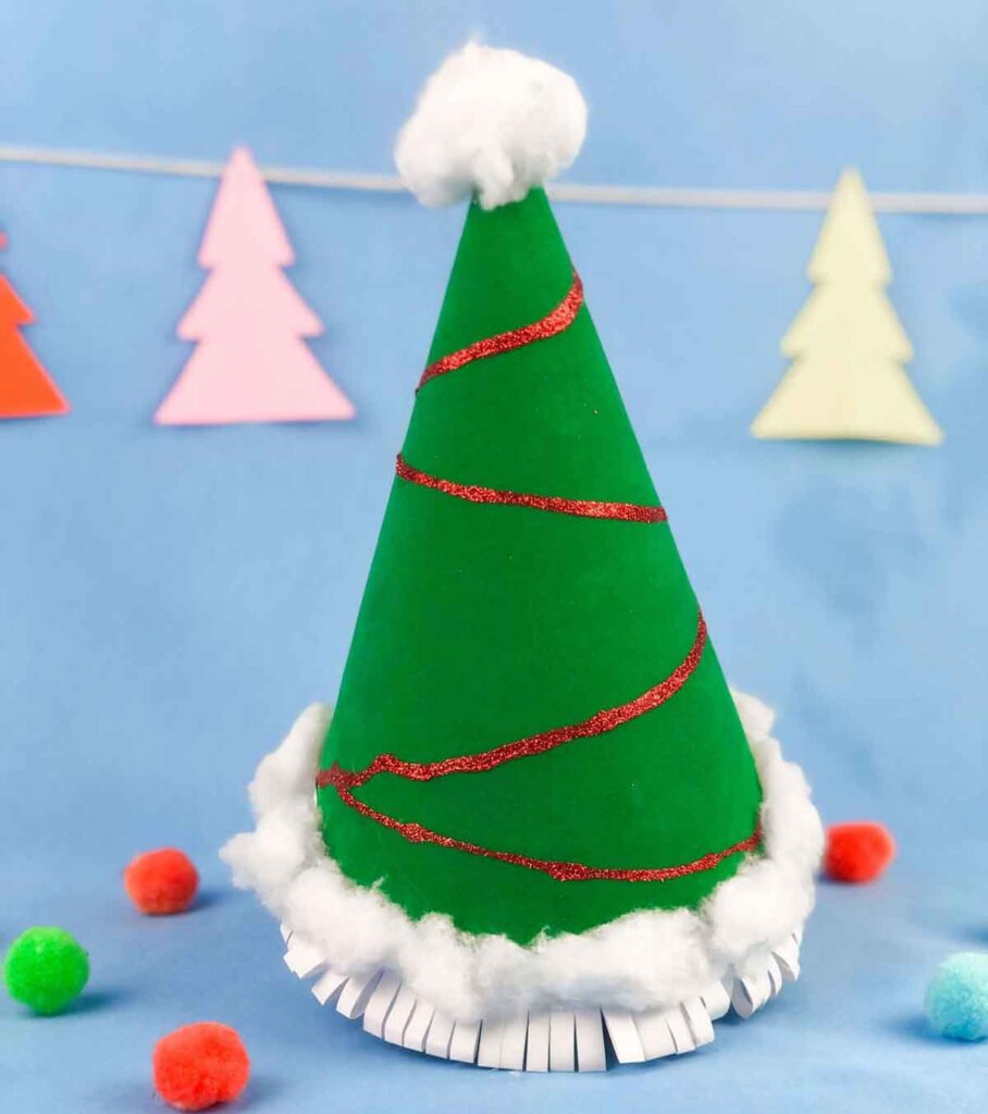 A green cone-shaped Christmas tree made from paper, decorated with a red glitter spiral and white cotton at the base, sits on a blue background. Colorful paper trees hang in the background, with red and green pom-poms nearby.