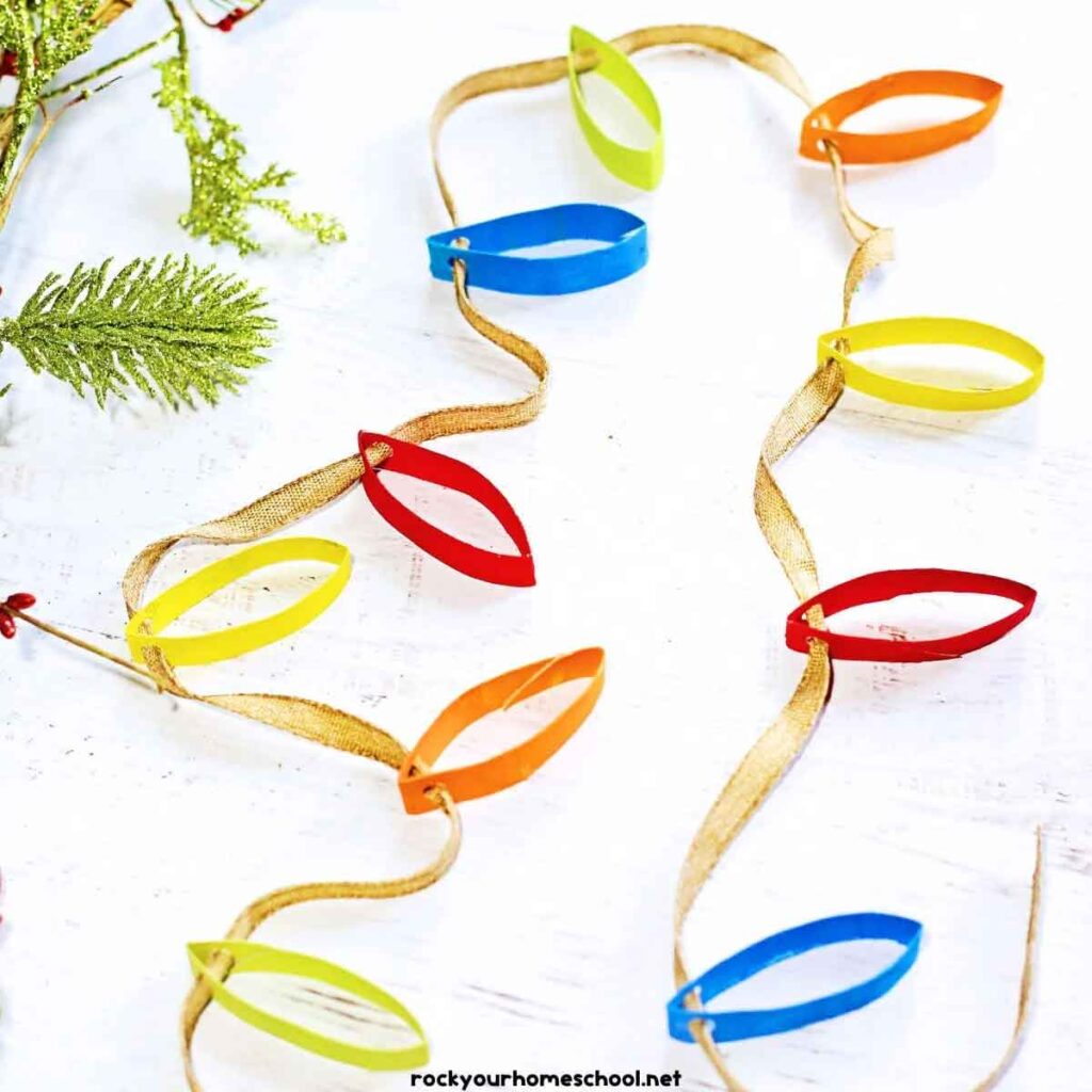 Colorful paper Christmas lights garland on a white surface makes a perfect project for christmas crafts for teens. The garland features alternating red, yellow, orange, blue, and green paper light shapes attached to a gold ribbon. Pine branches are visible in the background.