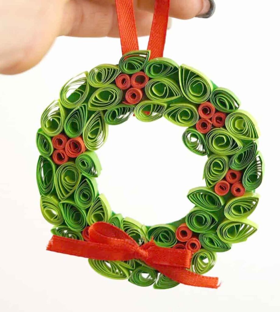 A hand holds a decorative wreath made of green and red quilled paper strips, perfect for Christmas crafts for teens. It features a red ribbon at the top and a charming red bow at the bottom.