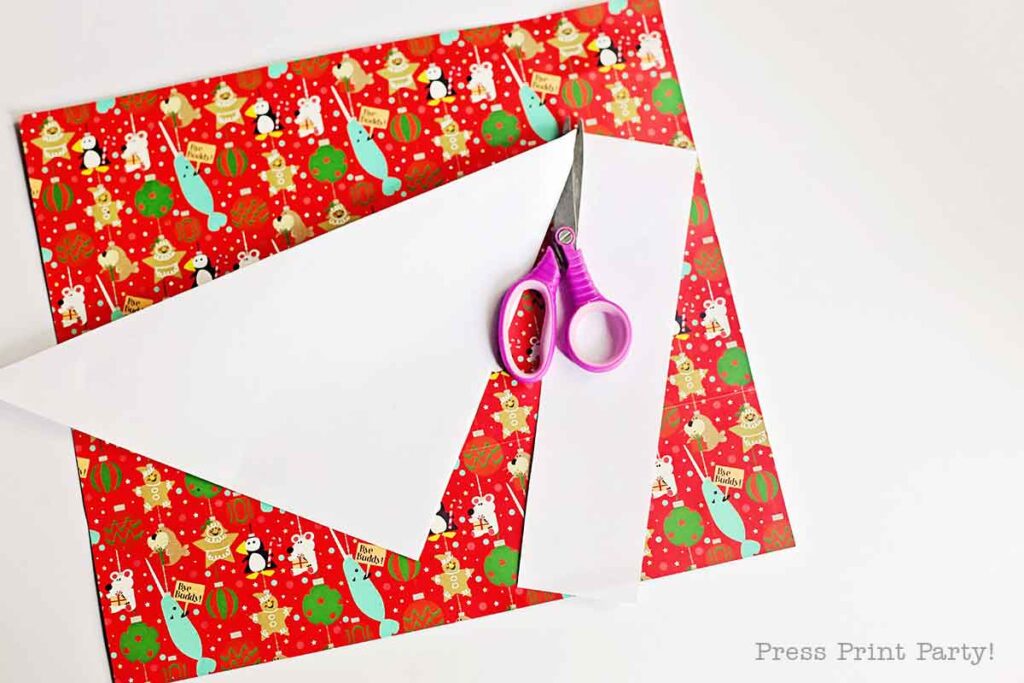 Colorful Christmas-themed wrapping paper with festive designs lies beneath a pair of pink scissors and a cut piece of white paper.