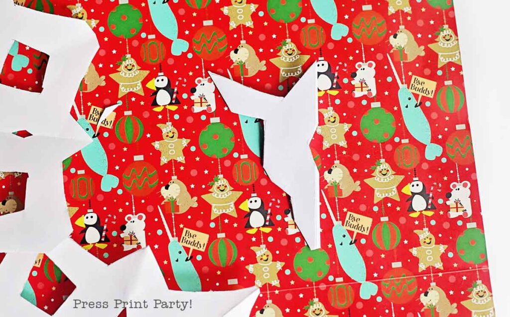 Colorful holiday wrapping paper featuring cartoon animals and festive decorations in red, green, and gold. Partially cut white paper snowflakes rest on top, with the phrase "Press Print Party!" visible in the corner.