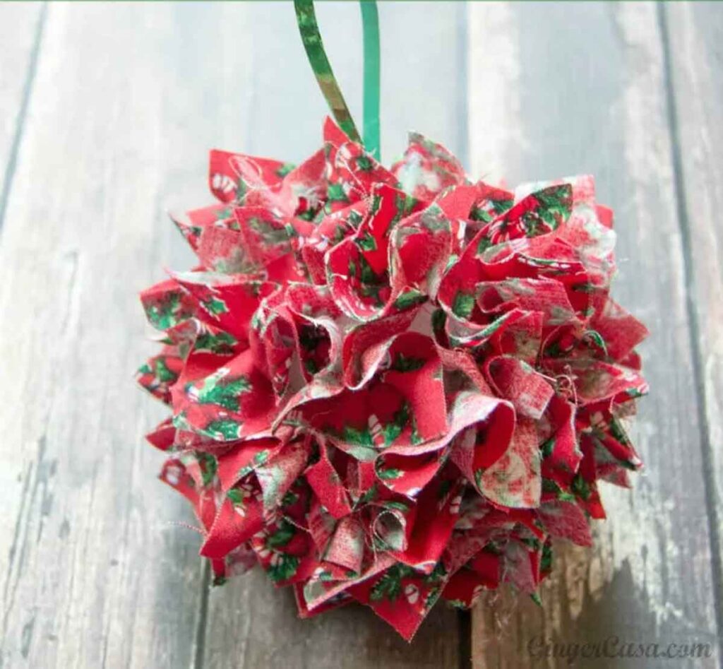 This holiday-themed ornament, perfect for Christmas crafts for teens, features red and green fabric squares gathered into a spherical shape. It hangs elegantly from a green ribbon against a rustic wooden background.