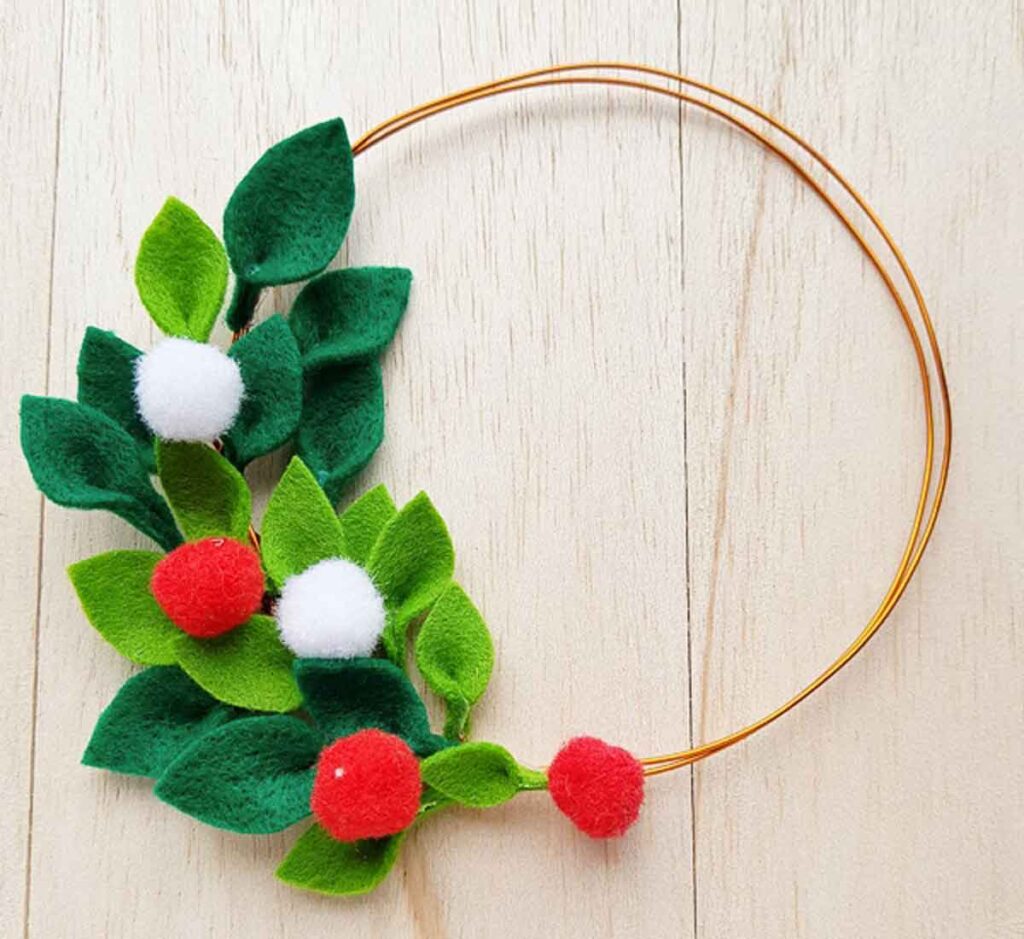 A circular wire wreath adorned with green felt leaves and red and white pom-poms sits elegantly on a light wooden surface, an ideal project for Christmas crafts for teens.