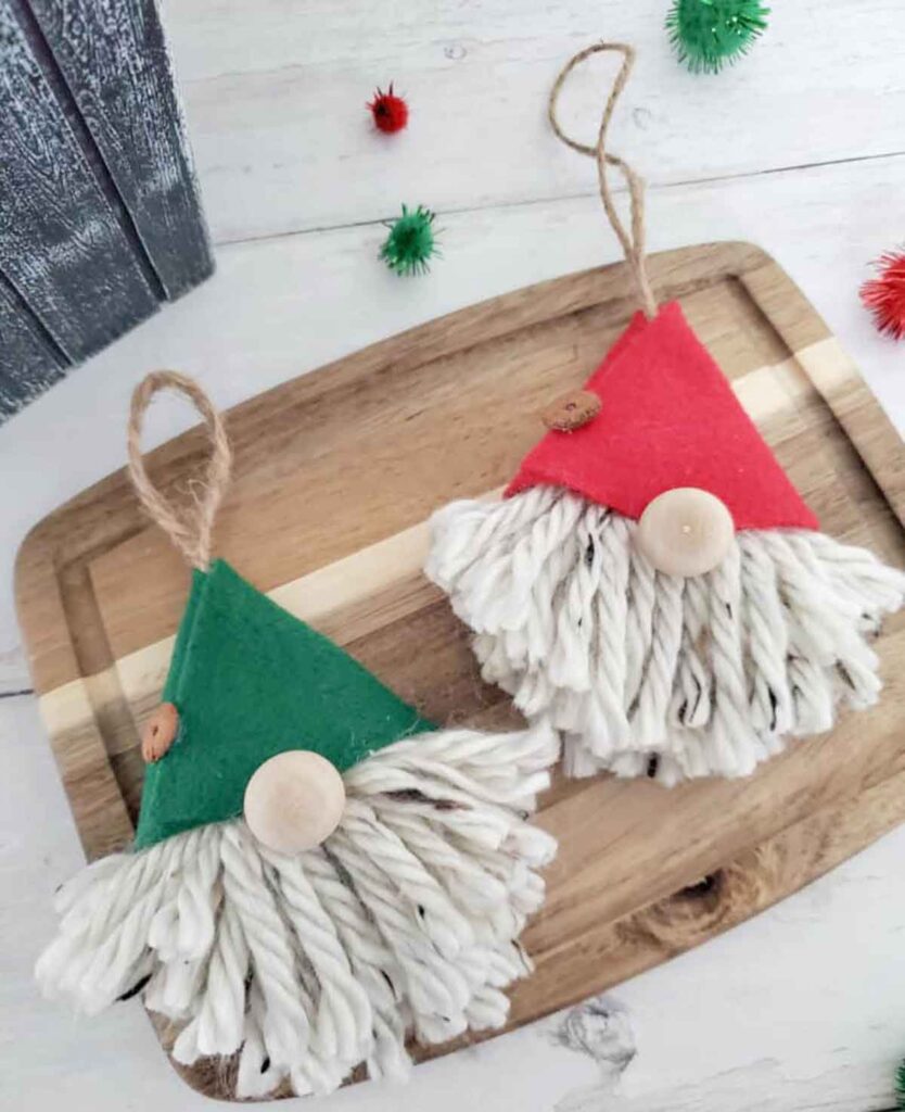 Two gnome ornaments with thick white yarn beards, one donning a green hat and the other a red, lie on a wooden tray—an ideal Christmas craft for teens. Both feature loops for hanging, while small, fuzzy craft balls are scattered around on the light wooden surface.