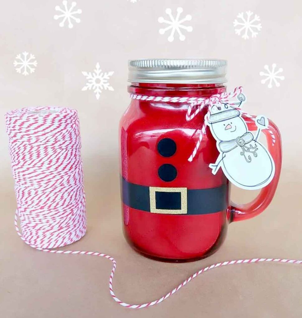 A mason jar filled with red liquid, decorated to resemble Santa's suit with a black belt and buttons, makes for a delightful Christmas craft for teens. It features a snowman tag attached with red and white twine, while a spool of matching twine rests nearby against snowflake designs.