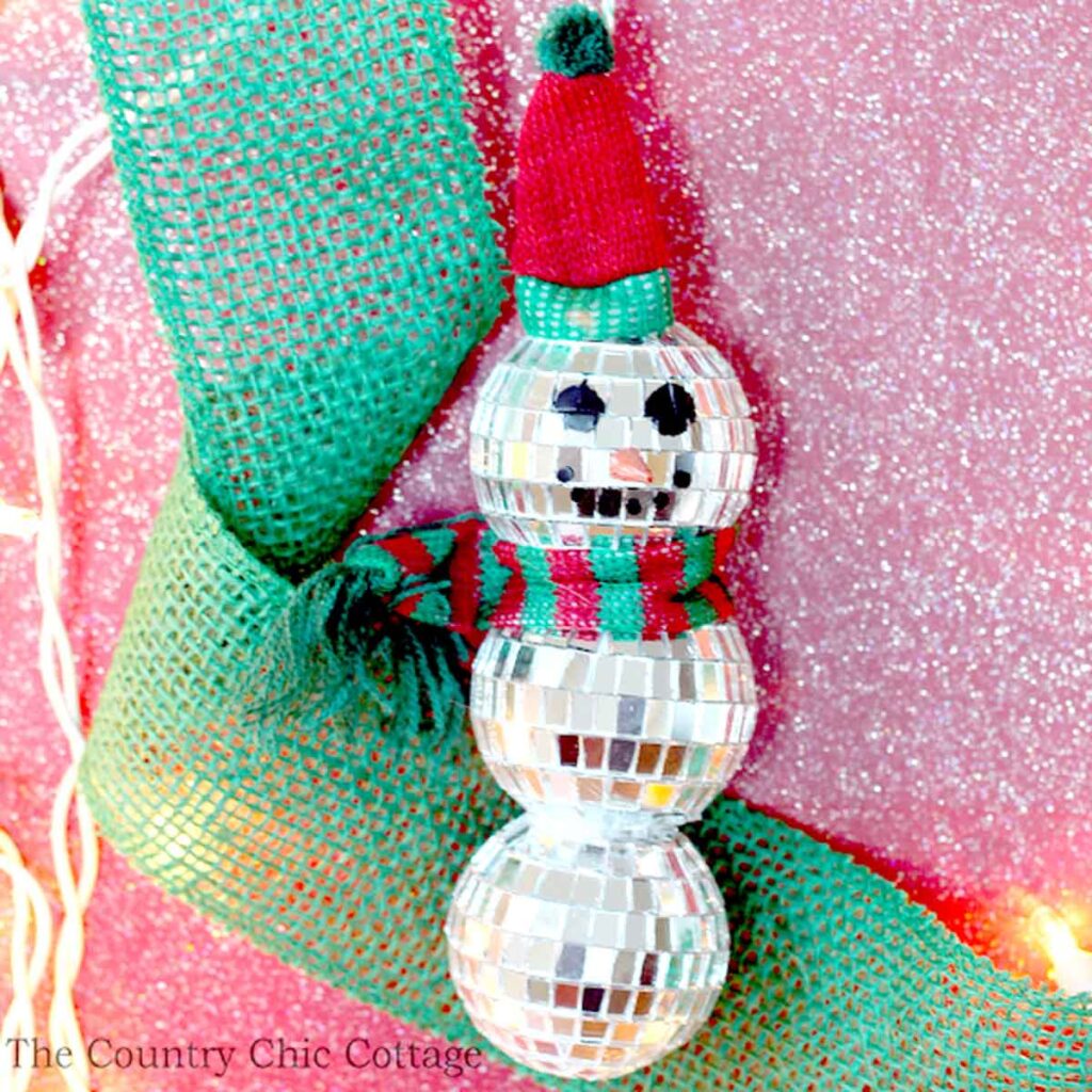 The festive snowman ornament, perfect for Christmas crafts for teens, is made of mirrored disco balls. It sports a red and green hat and scarf, hanging gracefully against a glittery pink background with green ribbon accents.