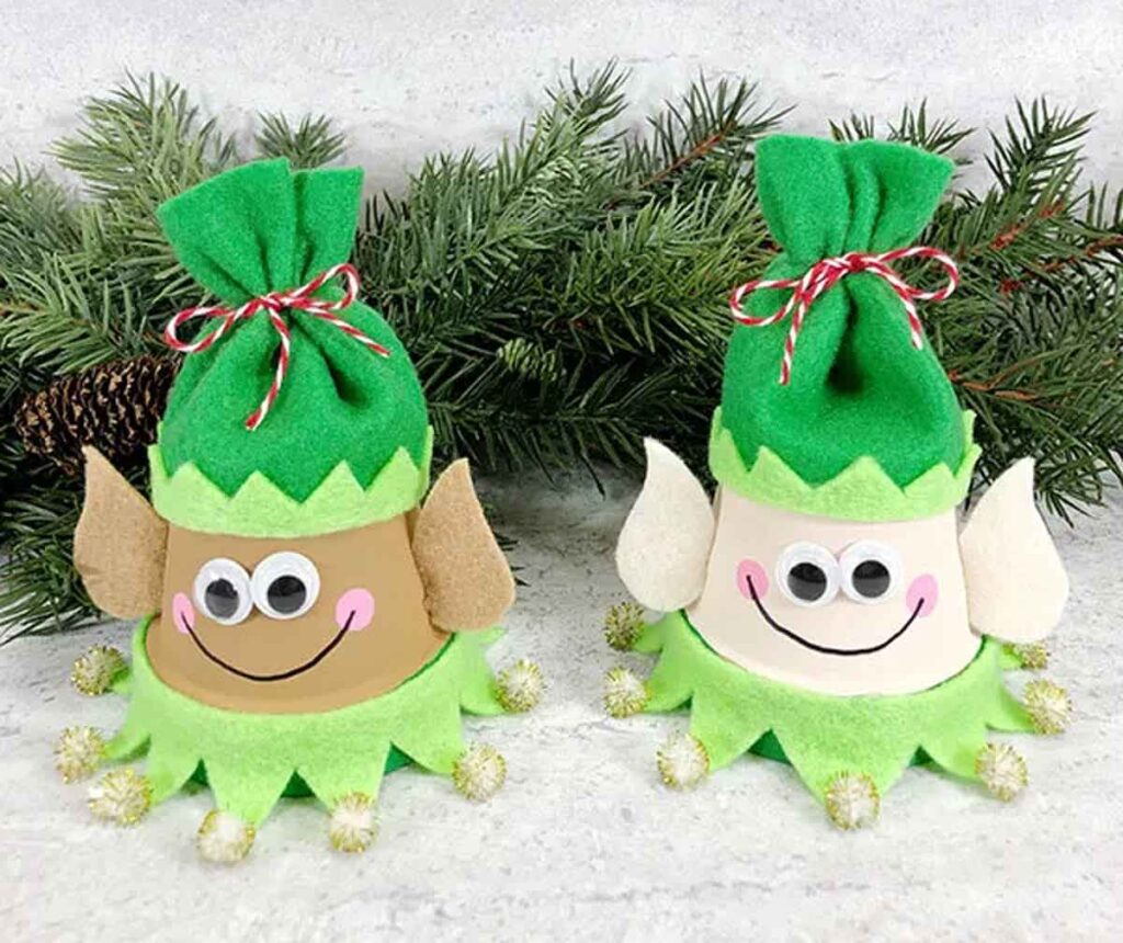 Two playful elf decorations with green hats, brown and white faces, and smiling expressions stand on a surface. Perfect for christmas crafts for teens, their base is adorned with jingle bells, while a pine branch adds a festive backdrop.