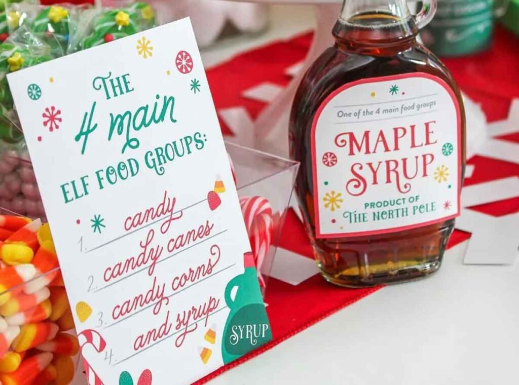 A festive holiday setup featuring a bottle of North Pole maple syrup next to a sign listing elf food groups: candy, candy canes, candy corn, and syrup. Bright colors and sweets create a cheerful atmosphere.