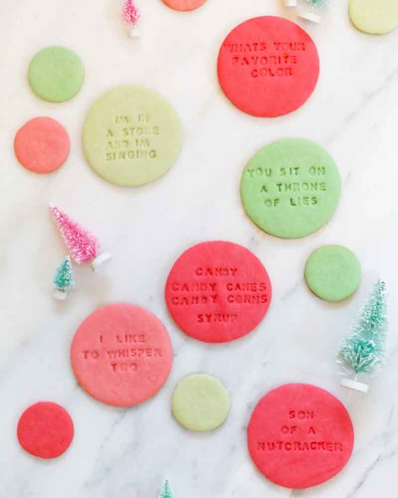 A marble surface with red and green round cookies featuring embossed text. Small decorative pink and green Christmas trees are scattered around the cookies.
