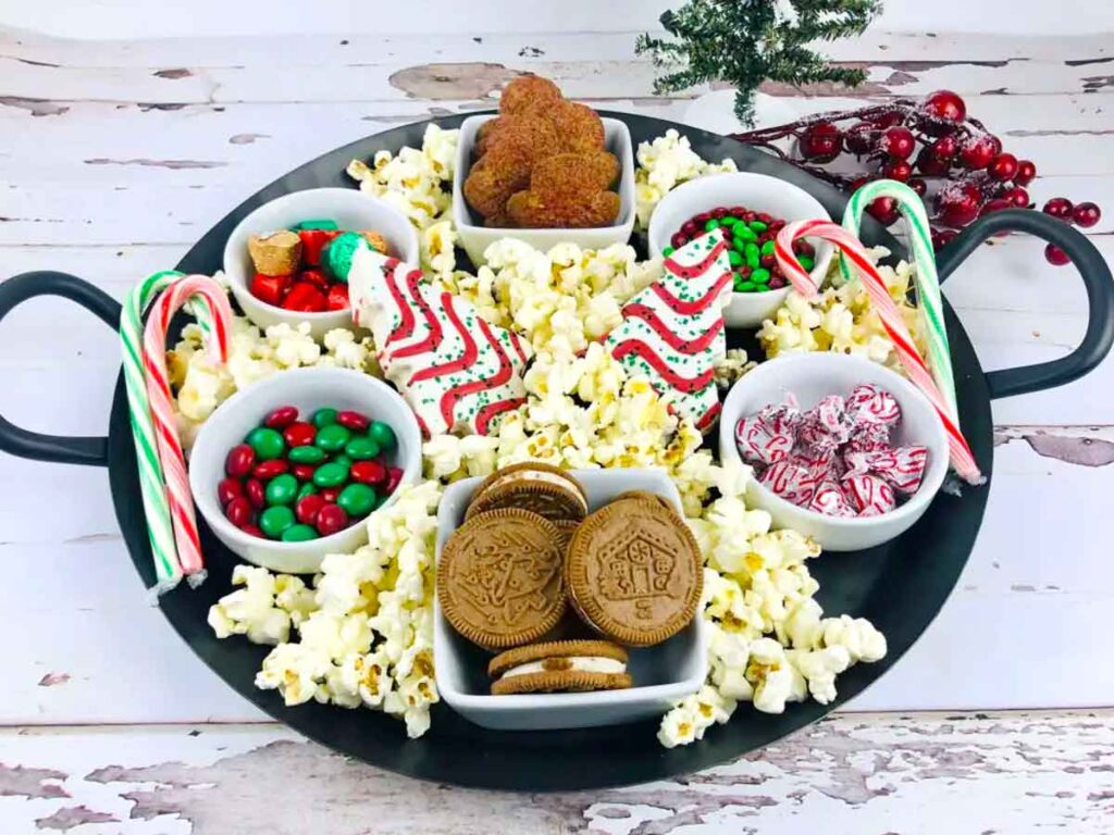 A festive tray with popcorn, gingerbread cookies, peppermint candies, chocolate cookies, and candy canes. Small bowls hold red and green candies. Christmas theme with a small tree and berries in the background.